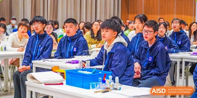 A male student in a blue jacket working with chemicals and lab equipment on a table during a science class at Wuxi Kuangyuan Bilingual School.