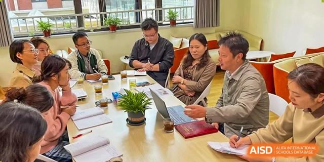 A small group of adults seated at a table for a meeting or discussion, with a laptop and potted plant visible, at Wuxi Kuangyuan Bilingual School.
