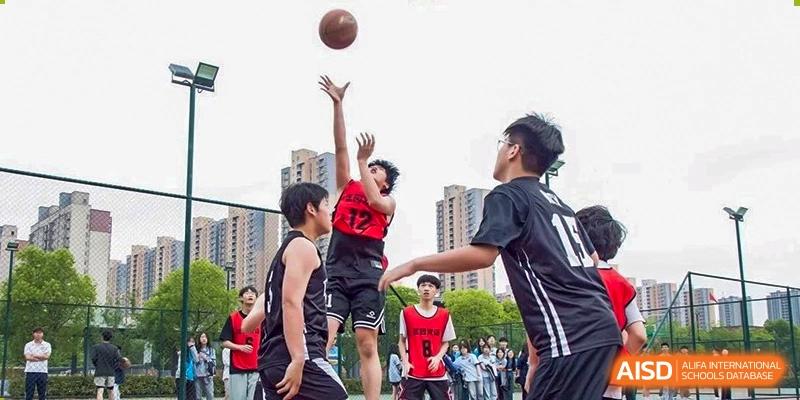 A student jumping up for a shot during an outdoor basketball game with teammates and onlookers at Wuxi Kuangyuan Bilingual School.
