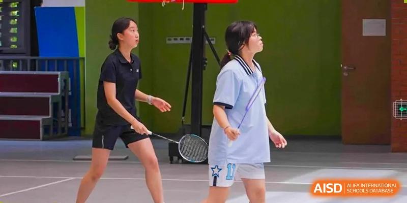 Two female students playing an indoor badminton match, one in a white and light blue uniform and one in a dark uniform, at Wuxi Kuangyuan Bilingual School.
