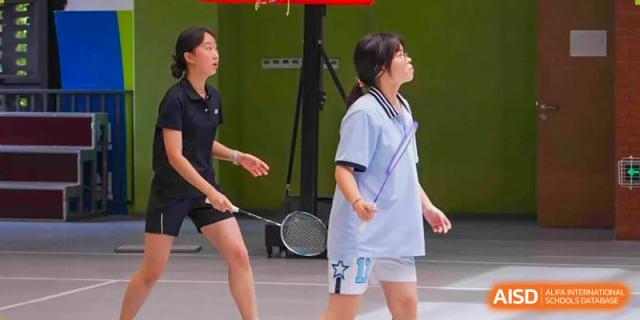 Two female students playing an indoor badminton match, one in a white and light blue uniform and one in a dark uniform, at Wuxi Kuangyuan Bilingual School.