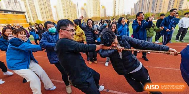 Adults intensely competing in a tug-of-war game on a sports field during a school event at Wuxi Kuangyuan Bilingual School.