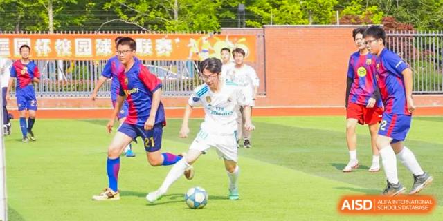 A male student in a white soccer uniform dribbling a ball during a game on a green turf field at Wuxi Kuangyuan Bilingual School.