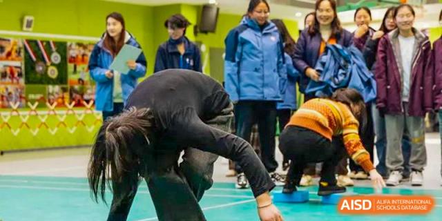 A student performing a gymnastic or athletic floor exercise indoors, with other students observing in the background, at Wuxi Kuangyuan Bilingual School.