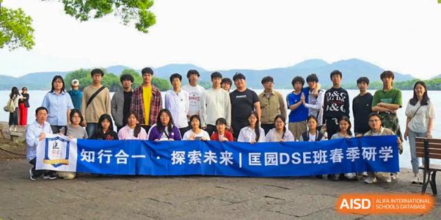 A group of students and teachers posing outdoors behind a banner that reads "探索未来 | 国DSB" next to a lake or body of water at Wuxi Kuangyuan Bilingual School.
