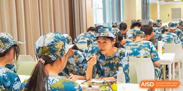 A male student in a patterned hat and uniform looking up while seated at a table during a school event or meal at Wuxi Kuangyuan Bilingual School.