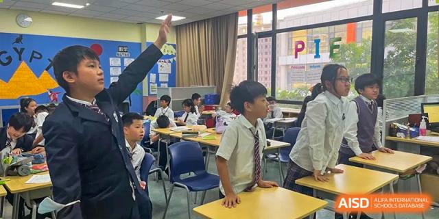 Student standing at his desk while a teacher points in a bright classroom at Wuxi United International School.