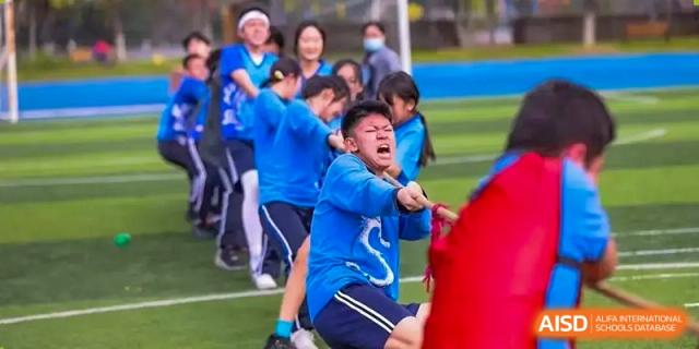 A student from Wuxi United International School, wearing a blue shirt, strains with effort while participating in a spirited tug-of-war game on a sports field, with teammates and cheering adults behind him.