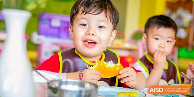 A smiling young child from Wuxi United International School wearing a bib and eating a snack or meal in a brightly colored setting.