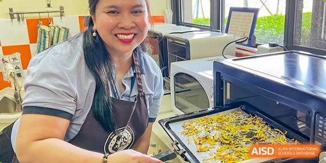 A smiling staff member from Wuxi United International School wearing an apron and holding a baking sheet of golden food next to an oven.