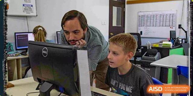 A Xi'an International School teacher leans over to help a male student who is working on a desktop computer.