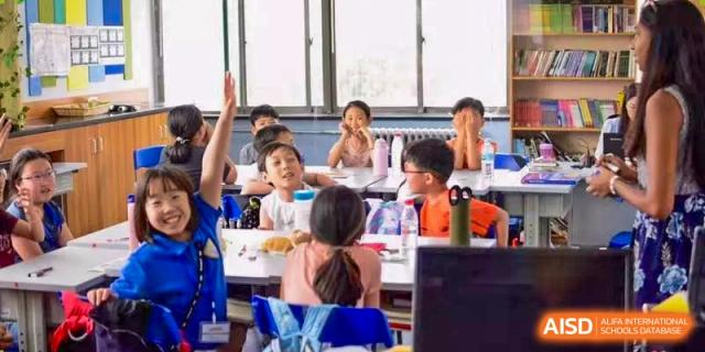 A group of enthusiastic Xi'an International School students seated at desks in a classroom, with several students raising their hands to answer a question.