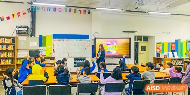 A Xi'an International School teacher instructs a class of young students seated at desks in front of a large, brightly lit display screen.
