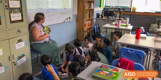 A group of young Xi'an International School students sitting on the floor in a bright classroom or library space, looking at the teacher.