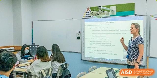 A female Xi'an International School student stands in front of a whiteboard in a classroom, giving a presentation or answering a question.