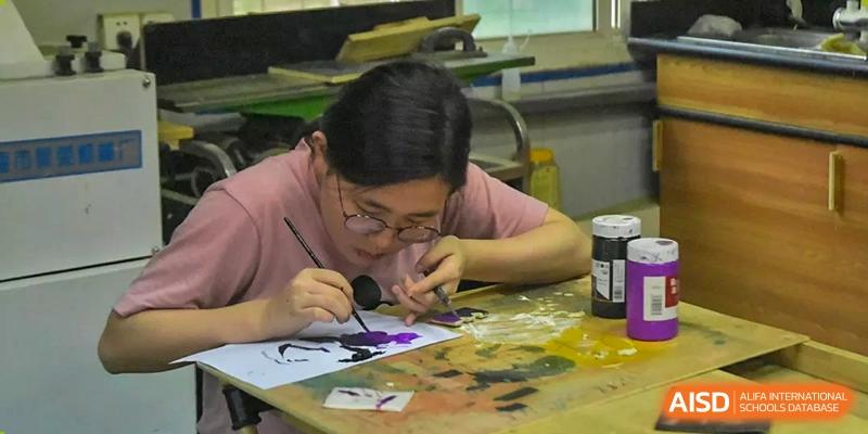 A Xi'an International School student wearing glasses and a pink shirt intently painting or crafting a small object in an art class or workshop.