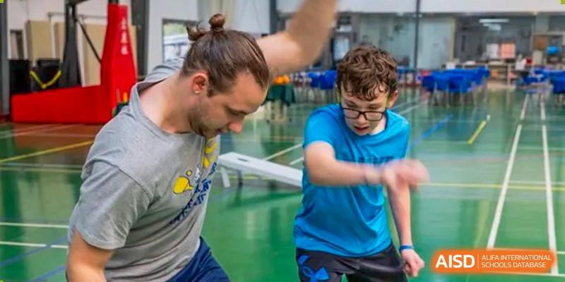 A Xi'an International School teacher with a bun playing a sport, possibly a variant of basketball, with a student in a blue shirt in an indoor gym.
