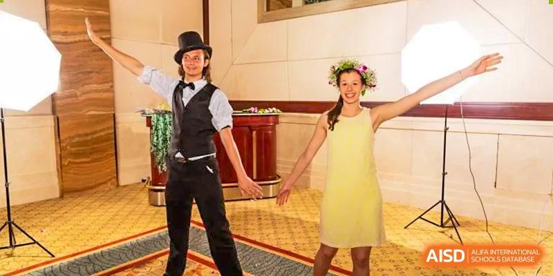 Two Xi'an International School students dressed for a formal or themed event, with one student in a vest and top hat and the other in a yellow dress and a flower crown.