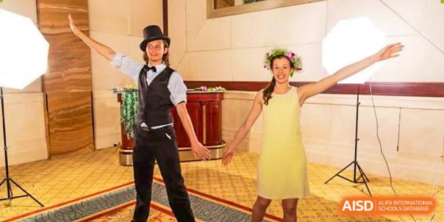 Two Xi'an International School students dressed for a formal or themed event, with one student in a vest and top hat and the other in a yellow dress and a flower crown.