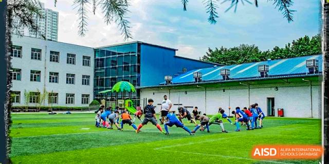 A wide shot of a Xi'an International School sports field showing a group of young students warming up or stretching in a line with a blue school building in the background.