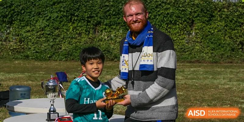 A smiling Xi'an International School coach in a scarf presenting a small golden trophy to a young male student in a green sports jersey.