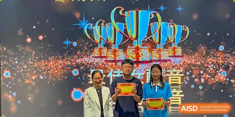 Students and a teacher from Xiamen Yingcai school holding certificates in front of a stage with trophies.