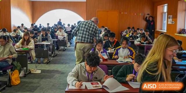 A group of students from Yantai Huasheng International School (YHIS) studying or writing in workbooks at tables during an event.