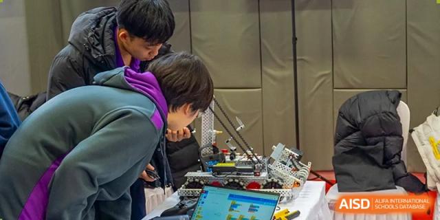 A student from Yantai Huasheng International School (YHIS) intently examining a complex robotics project or machine with a laptop nearby.
