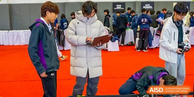 A student from Yantai Huasheng International School (YHIS) wearing a white puffer jacket and looking at a tablet or clipboard on a red carpet.
