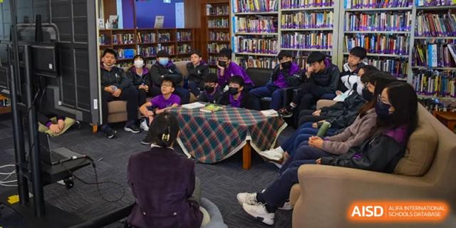 Students from Yantai Huasheng International School (YHIS) seated in a circular arrangement, reading or studying in a library surrounded by tall bookshelves.