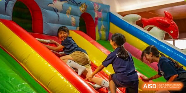 Students from Yantai Huasheng International School (YHIS) laughing while playing on a colorful inflatable bounce house slide indoors.