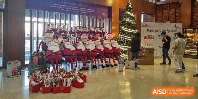 A group of people dressed as Santa Claus sitting in a row near a decorated Christmas tree inside the lobby or hall of Yantai Huasheng International School (YHIS).