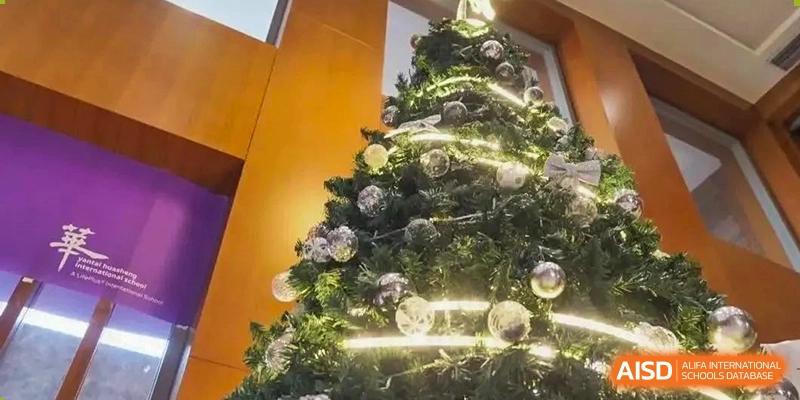 A close-up shot of a brightly lit and decorated Christmas tree with ornaments, likely located within the Yantai Huasheng International School (YHIS).