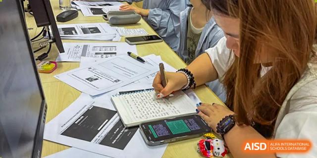 Close-up shot of a student taking notes in a spiral notebook and writing on papers spread across a desk during a class at BIBA (Tianjin International Bilingual Academy).