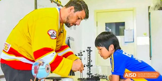 A male teacher, wearing a bright uniform or safety vest, guiding a young student using a microscope in a classroom at BIBA (Tianjin International Bilingual Academy).