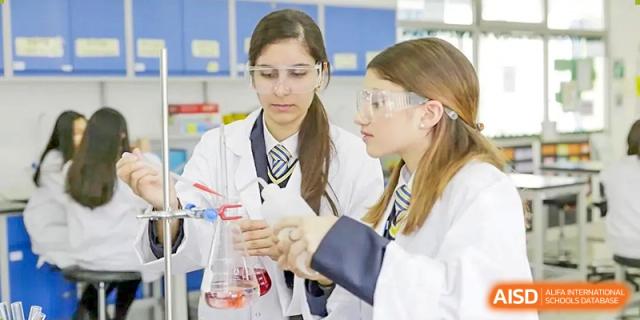 Two high school girls in lab coats and safety goggles working on a chemistry experiment, holding beakers in a science class at BIBA (Tianjin International Bilingual Academy).