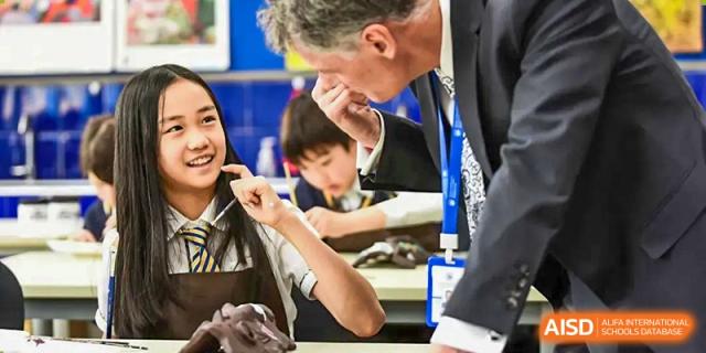 A male teacher in a suit leaning down to instruct or help a group of smiling, attentive students at their desks at BIBA (Tianjin International Bilingual Academy).