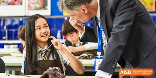 An adult male teacher in a suit interacting with and speaking to a smiling female student and a boy at a desk at BIBA (Tianjin International Bilingual Academy).