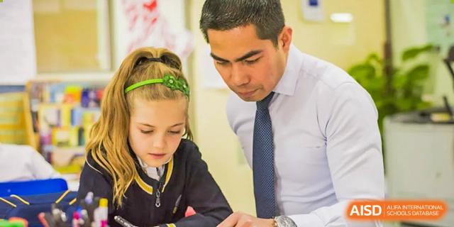 A male teacher in a white shirt and tie leaning over to help a young female student with her work in a classroom setting at BIBA (Tianjin International Bilingual Academy).