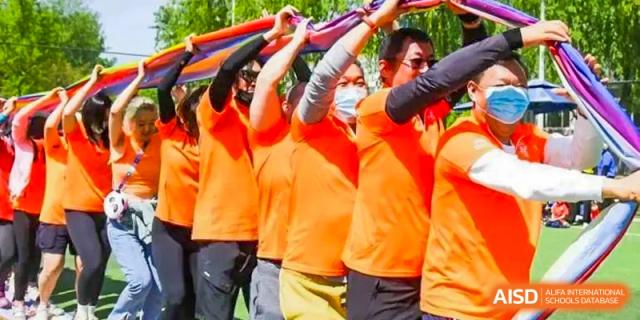 Group of people wearing orange shirts and sunglasses, some with face masks, holding colorful fabric overhead during an outdoor event at BIBA (Tianjin International Bilingual Academy).