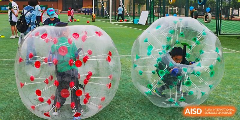 Students playing on a green field with large, transparent bubble soccer balls (bumper balls), showcasing outdoor sports activities at BIBA (Tianjin International Bilingual Academy).