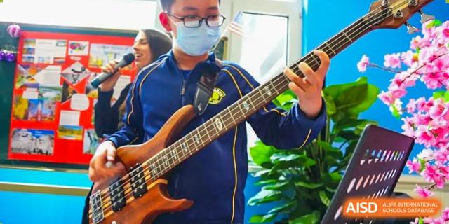 Student wearing a face mask and glasses playing a bass guitar during a music class or performance at BIBA (Tianjin International Bilingual Academy).