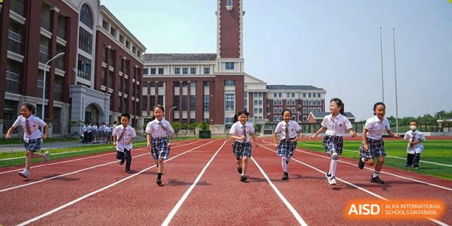 Students in uniform running on a red track with a large brick school building in the background at BIBA (Tianjin International Bilingual Academy).