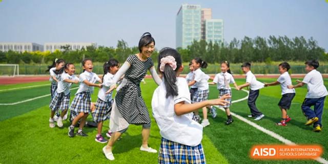 An enthusiastic teacher playing and dancing with a group of cheerful students in uniform on a grassy outdoor field at BIBA (Tianjin International Bilingual Academy).