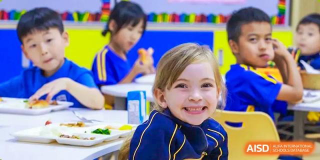 Close-up of a happy young student with blonde hair smiling while eating a meal in the dining area or classroom at BIBA (Tianjin International Bilingual Academy).