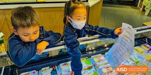 A young student wearing a protective face mask reaching into a display case to select a snack or pastry in the school cafeteria at BIBA (Tianjin International Bilingual Academy).