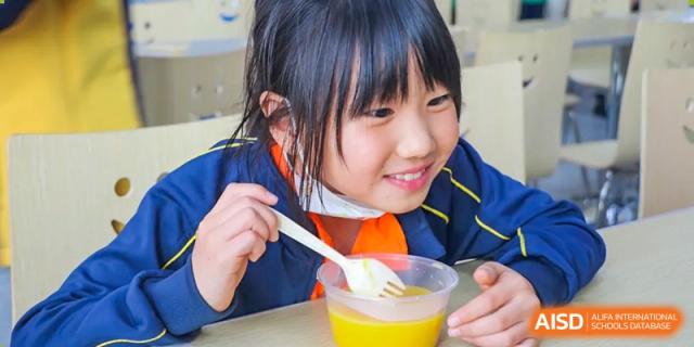 A female student with dark hair, wearing an orange shirt and school jacket, enjoying a bowl of food with a spoon during lunchtime at BIBA (Tianjin International Bilingual Academy).