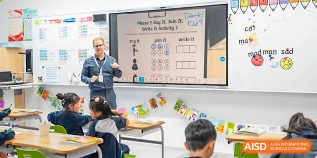 A male teacher from Dingwen Academy Hangzhou teaching a lesson in a classroom with a digital presentation on a large screen.