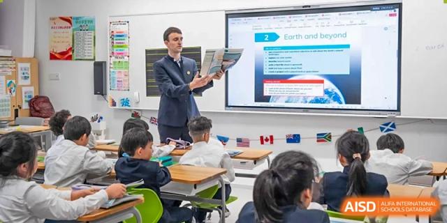 A male teacher from Dingwen Academy Hangzhou holding a textbook and instructing students seated at their desks in a modern classroom.