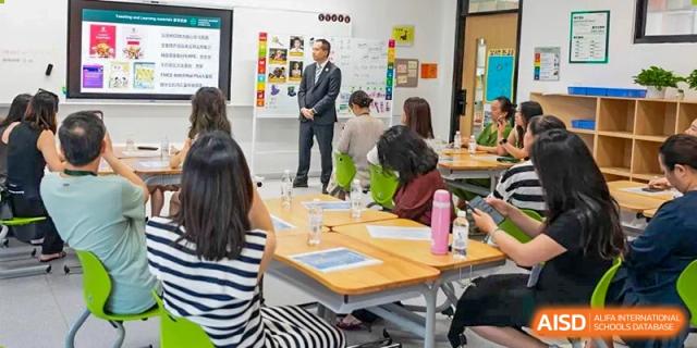 An adult from Dingwen Academy Hangzhou addressing a group of students and adults seated at tables in a bright, modern classroom.